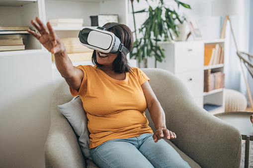 Mature black woman wearing virtual reality headset while sitting in living room at home.