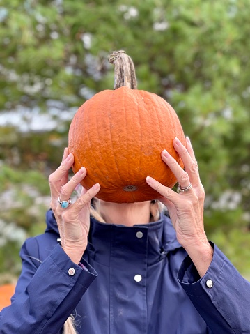Active senior woman having fun holds pumpkin up above her neck covering her face and hair outdoors in autumn season in Wisconsin.  Portrait