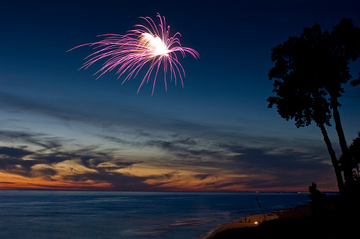 Vivid, colorful fireworks are being launched on the beach, casting interesting reflections on the water and beach below. Trees are darkly silhouetted against a vivid sunset.