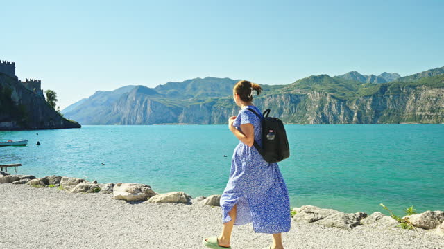 SLO MO Female Tourist With Backpack Walking On Shore Of Lake Garda near Malcesine Castle On Sunny Day