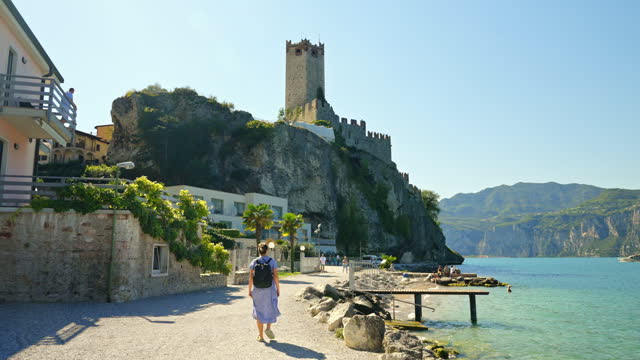SLO MO Young Female Backpacker Walking on Lakeside Of Lake Garda Leading towards Scaliger Castle under Blue Sky