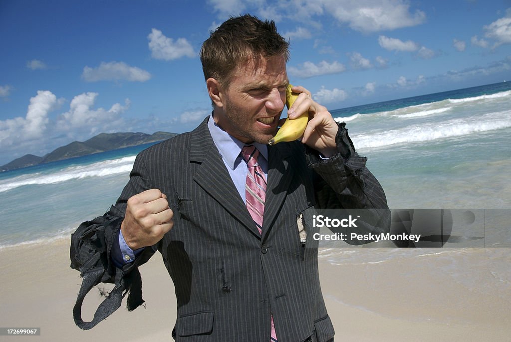 Frustrated Castaway Businessman Talks on Mobile Banana Castaway businessman stands with an angry expression and clenched fist talking on his banana phone Torn Stock Photo