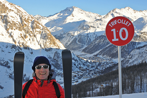 Senior skier standing with pride next to ski slope sign above Val d'Is