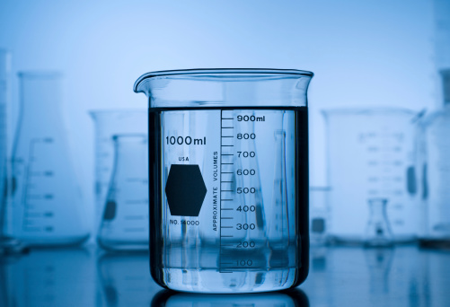 An Beaker filled with  fluid with a stock of laboratory glass in the background.