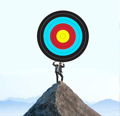 A man stands on top of a mountain peak and struggles to lift a large target above his head.