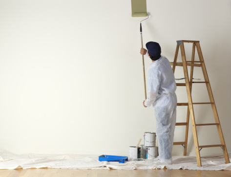 A man dressed in coveralls, uses a paint roller on a long stick  to paint a large wall. A nearby ladder waits in case the painter needs to paint the higher points on the wall.  Several paint cans sit on the painter's cloth draped over a hardwood surface. A large expanse of empty wall is available for copy.