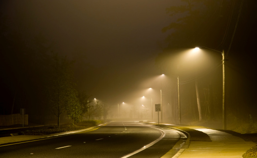 A light fog hangs over a road illuminated by streetlight at night. The road and sidewalks are slightly wet.