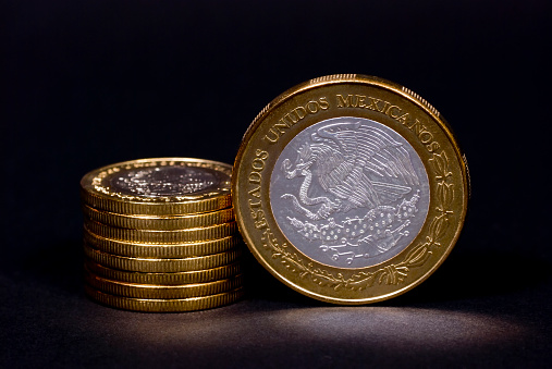 A $100 coin with a stack of $10 (mexican) pesos coins in a dark texturized background.