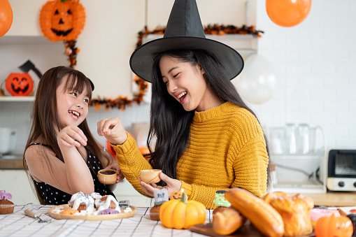 A happy and joyful young Asian girl and her mom in Halloween costumes are laughing, having fun, enjoying decorating Halloween cupcakes in the kitchen together. Spooky season, family time