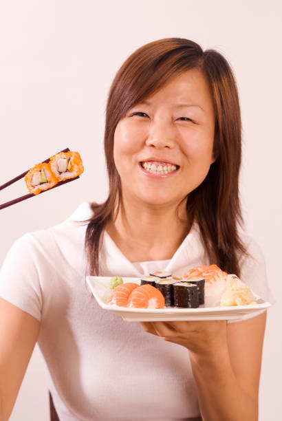 Young Asian Girl With 2 Sushi Maki Between Chopsticks Young Asian woman with sushi mix plate and 2 Inside Out at StAbchen kids eating tuna stock pictures, royalty-free photos & images