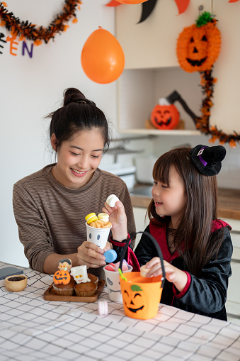 A happy and cute young Asian girl in a Halloween costume enjoys preparing Halloween desserts for trick-or-treat with her mom in the kitchen, celebrating Halloween at home together.