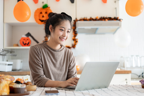 A beautiful and happy Asian woman is using her laptop, working on her computer at a kitchen table during the Halloween season at home.