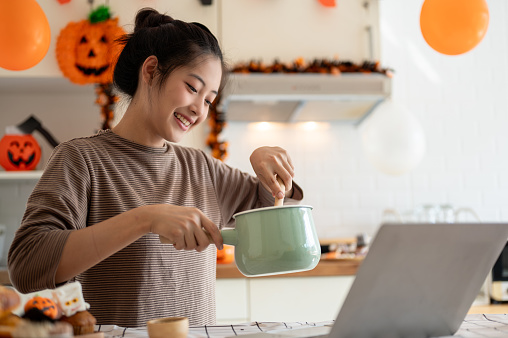 A cheerful and happy young Asian woman is learning Halloween cupcake recipes online and making Halloween cupcakes in the kitchen. Leisure concept