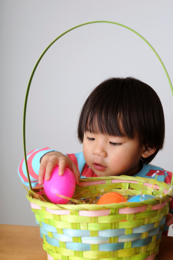 A two-year old girl reaches into a colorful Easter basket on a table and grabs a plastic Easter egg.