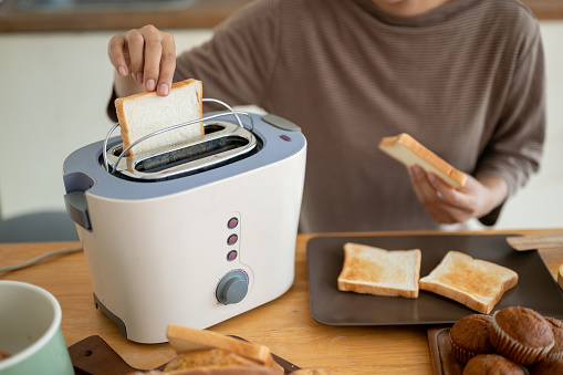 Close-up image of a beautiful young Asian woman is making tasty toasts with a toaster in the kitchen, preparing her breakfast. Cooking concept