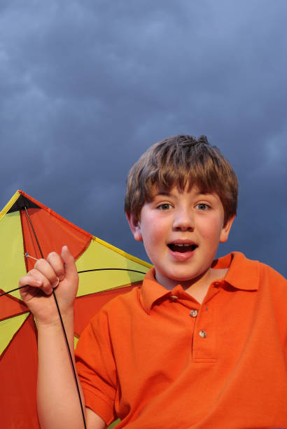 A cheerful young Asian boy in a blue cap adjusts his hat outdoors. Casual grey shirt, bright light, and a relaxed, playful mood emphasize youth, happiness, and outdoor fun.