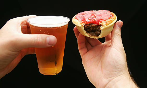 Beer and Meat Pie with Sauce A beer in a plastic cup and a meat pie covered in sauce with a bite out of it. Standard meal at the footy meat pie stock pictures, royalty-free photos & images