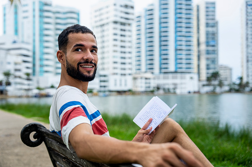 Mid-shot of relaxed Latin-American male tourist reading on a bench at Bahia El Laguito, Cartagena, Colombia