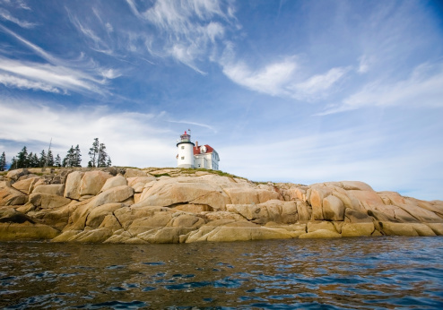 beautiful lighthouse with red roof near pines on large rocks surrounded by water and a sweeping blue sky