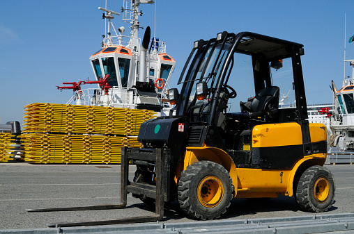 Forklift ready to load stack of wooden boards,tugboat in the background.