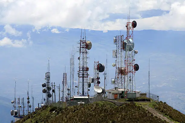 Photo of several communication antennas and tours in Quito, Ecuador. The communication antennas are located on the top of a mountain summit. The sky is very clouded and it is a grey day. There is a mountain range in the foggy background. 