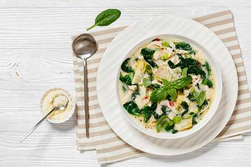 chicken spinach artichoke creamy soup in white bowl on white textured wooden table with spoon, american cuisine, horizontal view from above, flat lay, free space
