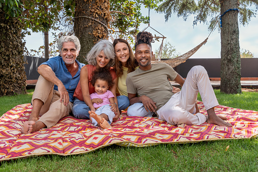 A happy family, including grandparents, parents with diverse backgrounds, and a child, sit together on a blanket in the garden. The African father sports rasta hair while they all smile for the camera