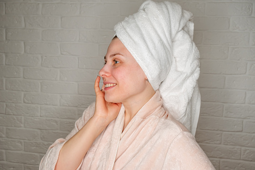 Natural beauty, home care for problem skin. Cute woman in white coat and towel on her head makes herself a makeup. Woman in bathroom wrapped in towel in bathrobe making lifting and massaging her skin.