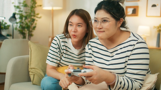 Happy young attractive Asian woman hand holding wireless controller joystick enjoy playing console game with friend or sister  feeling cheerful fun and excited sit on couch at cozy home living room.