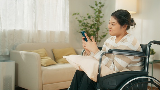 Portrait of Beautiful Asian woman in wheelchair using mobile phone browsing the internet, online shopping, texting a message while sitting in the living room at home