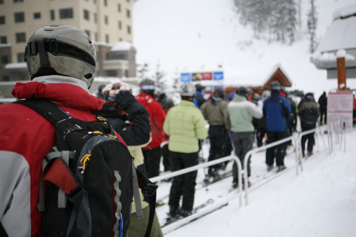 Skiers and snowboarders waiting in line for chair lift with falling snow.