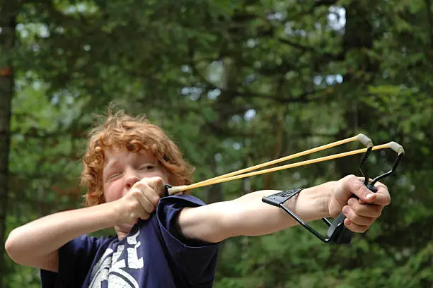 Nine-year-old boy is ready to fire his slingshot at some pop cans tied up in the trees.You can feel the tension! Focus is on his left hand.