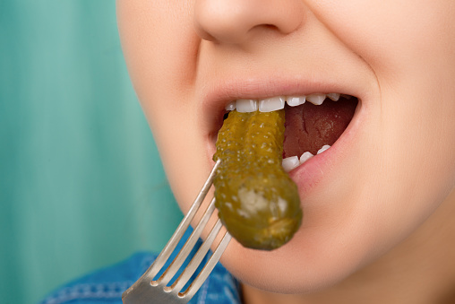 Close-up of a pretty Caucasian woman's mouth biting a pickled cucumber. Front three-quarter view. Low angle view. Indoors.