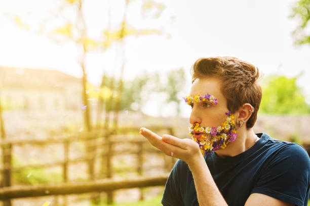 Surrounded by nature, a young man with facial hair made of spring blossoms releases petals into the air. This evocative scene beautifully captures the essence of life and poetic romance