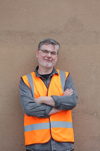 Portrait of a mature male volunteer wearing an orange reflective vest standing outdoors with arms crossed against brown wall.