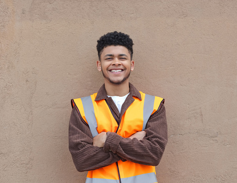 Portrait of a gen z young male volunteer wearing an orange reflective vest standing outdoors with arms crossed against brown wall.