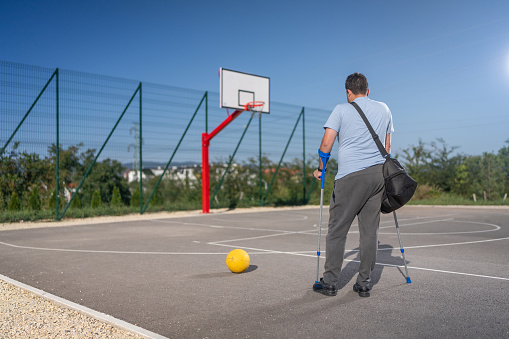 Man with crutches standing on basketball court outdoors.