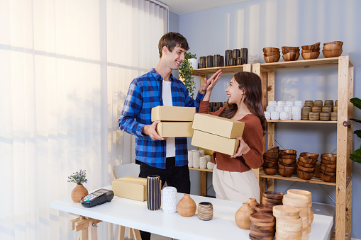 Success couple of Caucasian male and Asian female sell vase products online live streaming at home. Standing, high five, holding parcels for sending by order to the customers. Shopping online concept.