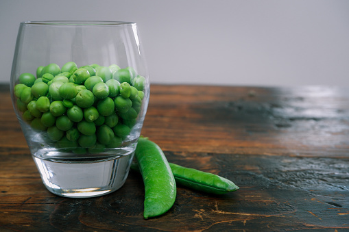Green peas in glass bowl. fresh pea in the pod with green leaves. green peas on a brown wodden table. Shelled peas in a glass with the pods on the side