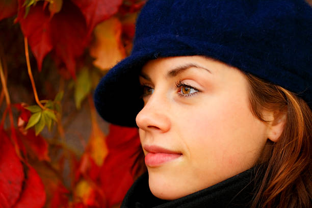 Gorgeous young woman standing beside a wall of red autumn leaves. She is wearing a black winter coat and a blue wool hat and looking to the side.