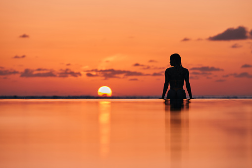 Rear view of silhouette of a woman getting out of infinity pool during summer sunset on the beach. Copy space.
