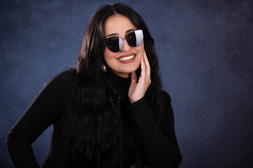 Young lady studio portrait over blue background. She looks at the camera while putting her hand on her chin has a big smile. She wears a black sweater and black sunglasses.