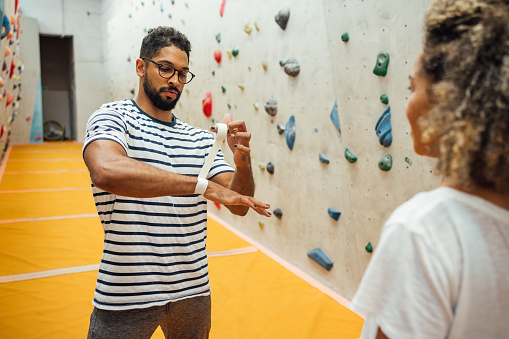 An over-the-shoulder shot of an adult male winding athletic tape around his wrist ready for a climbing session at a climbing centre in Byker, Newcastle upon Tyne. He is standing opposite his friend whilst focusing on the task at hand. They are standing on a safety mat beside one of the climbing walls which features multicoloured hand and foot-holds of varying sizes.