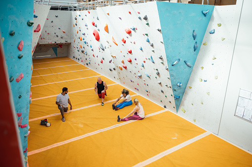 An elevated shot of a small group of people warming up before a climbing session in a climbing centre in Byker, Newcastle upon Tyne. They are all warming up and stretching their muscles. The floor is covered by a safety mat and the climbing walls feature multicoloured hand and foot-holds of varying sizes.