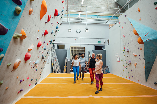 A shot of a small group of people walking across a safety mat towards a climbing wall in a climbing centre in Byker, Newcastle upon Tyne. They are all wearing sports clothing ready for their climbing session. The climbing wall features multicoloured hand and foot-holds of varying sizes.
