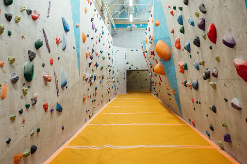 A shot of an empty climbing wall in a climbing centre in Byker, Newcastle Upon Tyne. On the walls there are a variety of colourful hand and foot-holds of varying sizes. The yellow safety flooring is visible to help people when they descend. At the far end is an archway leading to another area of the climbing centre.