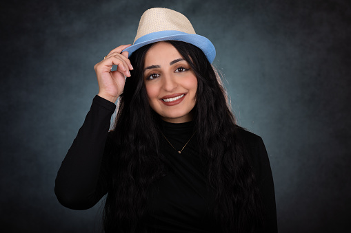 Young lady studio portrait over green background. She looks at the camera and has a big smile while saluting with her hand on a hat. She wears a black sweater and has long black hair