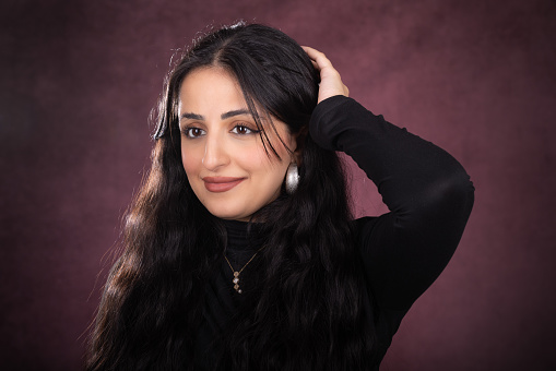 Young lady studio portrait over red background. She has a small smile and rests her hand on the back of her head like she is thinking. She is wearing a black sweater and has long black hair.