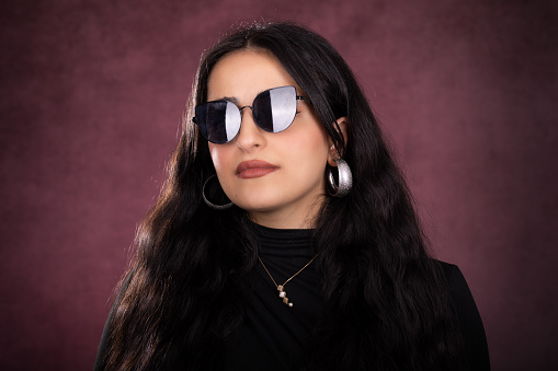 Young lady studio portrait over red background. She is serious, looking away and wears sunglasses. She wears a black sweater and has long black hair.