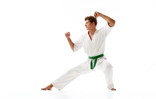 Young guy, karate sportsman in white kimono and green belt standing in pose to fight isolated on white studio background. Concept of martial arts, combat sport, energy, strength, health. Ad